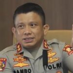 Male police officer in uniform seated at a desk, raising his index finger as if making a point.