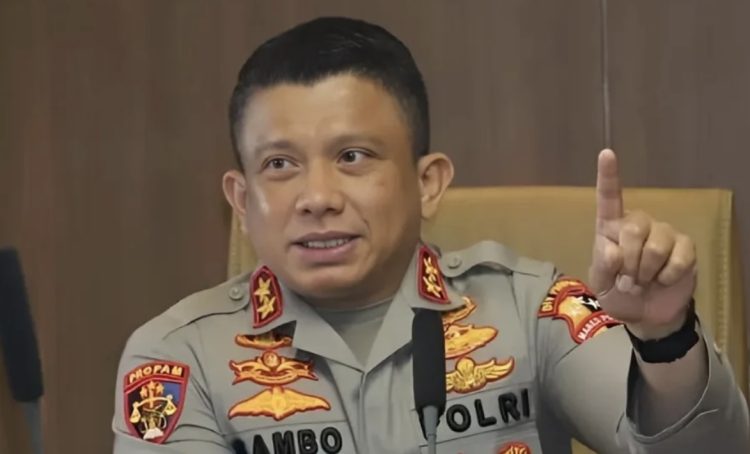 Male police officer in uniform seated at a desk, raising his index finger as if making a point.