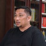 Man in a black shirt sits in a home library, speaking, with bookshelves behind him