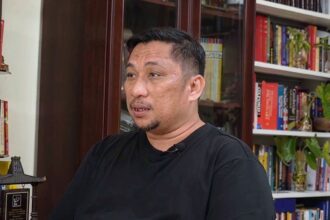 Man in a black shirt sits in a home library, speaking, with bookshelves behind him
