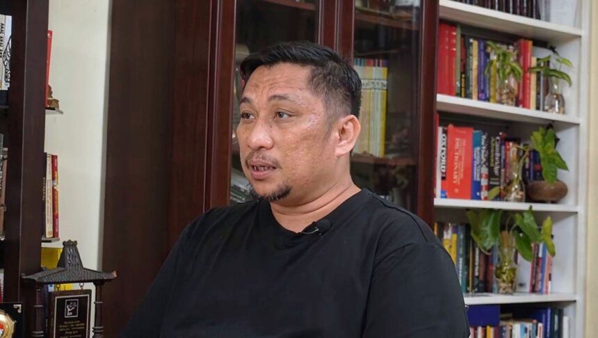 Man in a black shirt sits in a home library, speaking, with bookshelves behind him