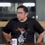 Man with glasses in a black T-shirt speaks into a microphone at a podcast recording setup with two mics on stands in front of him.