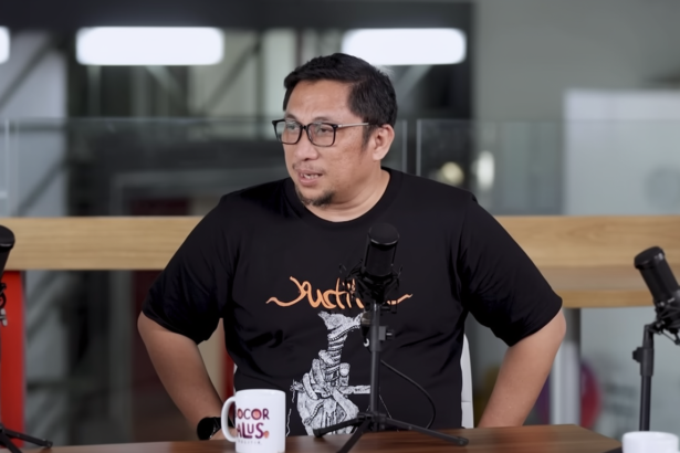 Man with glasses in a black T-shirt speaks into a microphone at a podcast recording setup with two mics on stands in front of him.