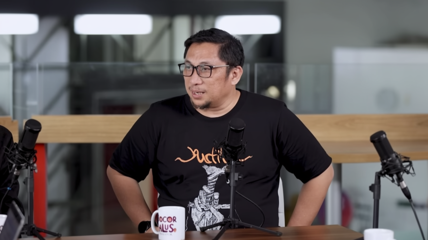 Man with glasses in a black T-shirt speaks into a microphone at a podcast recording setup with two mics on stands in front of him.