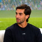 Man with dark hair and stubble wearing a black athletic shirt sits on a white sofa with a blue cushion, appearing to be in an interview setting.