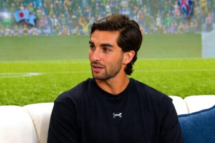 Man with dark hair and stubble wearing a black athletic shirt sits on a white sofa with a blue cushion, appearing to be in an interview setting.