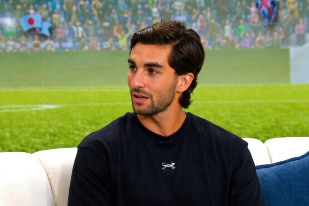 Man with dark hair and stubble wearing a black athletic shirt sits on a white sofa with a blue cushion, appearing to be in an interview setting.