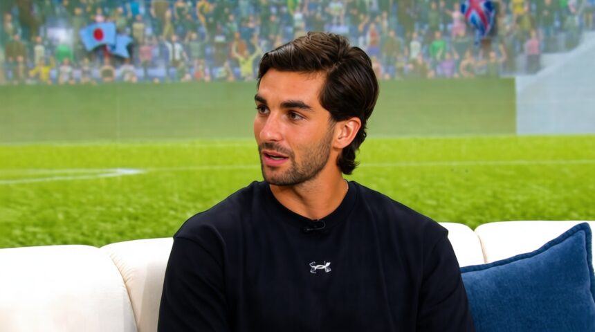 Man with dark hair and stubble wearing a black athletic shirt sits on a white sofa with a blue cushion, appearing to be in an interview setting.