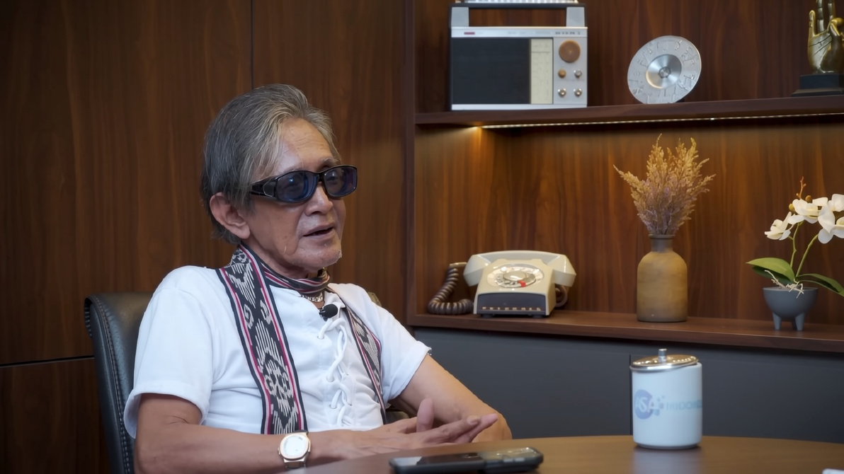 Older man with sunglasses sits at a desk in a wood-paneled office, talking during an interview.