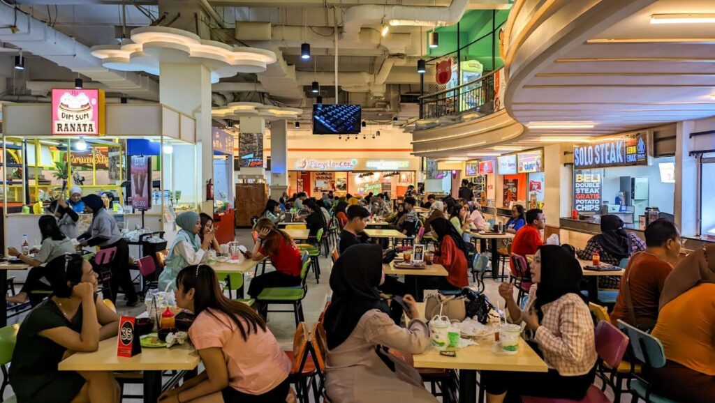 A busy indoor food court with many people seated at colorful tables, eating and chatting under bright ceiling lights.