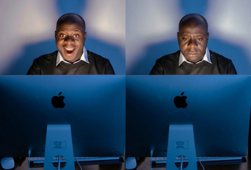 Two men sit side by side at blue Apple computer monitors; left man smiles broadly, right man looks down in a somber mood.