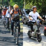 A large group of cyclists wearing helmets ride together on a sunny street; front riders in casual shirts and colorful jerseys.