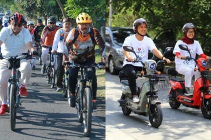 A large group of cyclists wearing helmets ride together on a sunny street; front riders in casual shirts and colorful jerseys.