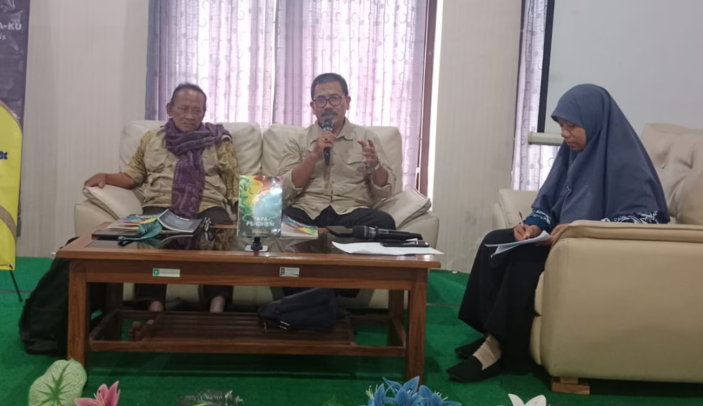 Three panelists sit on beige sofas during a discussion; the middle man speaks into a microphone while the others listen, and a woman on the right takes notes.