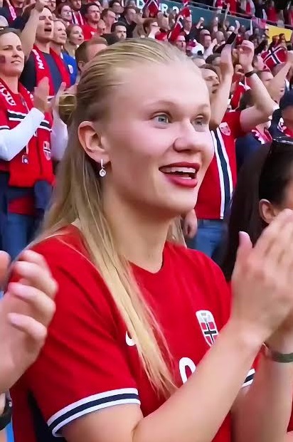 Blonde woman in a red Norway football jersey clapping in a crowded stadium.