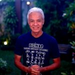 Smiling older man with white hair, wearing a Bisindo T-shirt, standing in a garden at dusk.