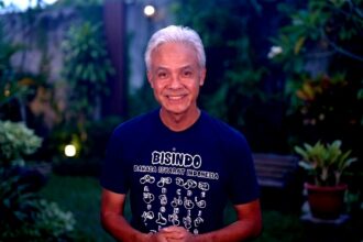 Smiling older man with white hair, wearing a Bisindo T-shirt, standing in a garden at dusk.