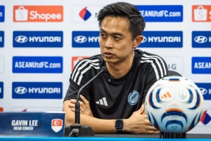 Press conference: man in black athletic shirt, nameplate reads Gavin Lee, head coach, at a football media briefing with logos in background
