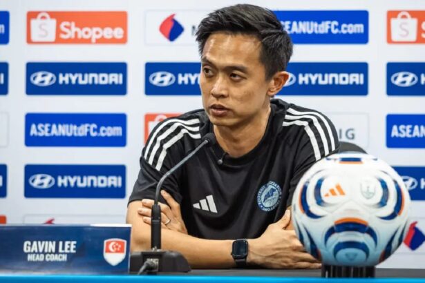 Press conference: man in black athletic shirt, nameplate reads Gavin Lee, head coach, at a football media briefing with logos in background