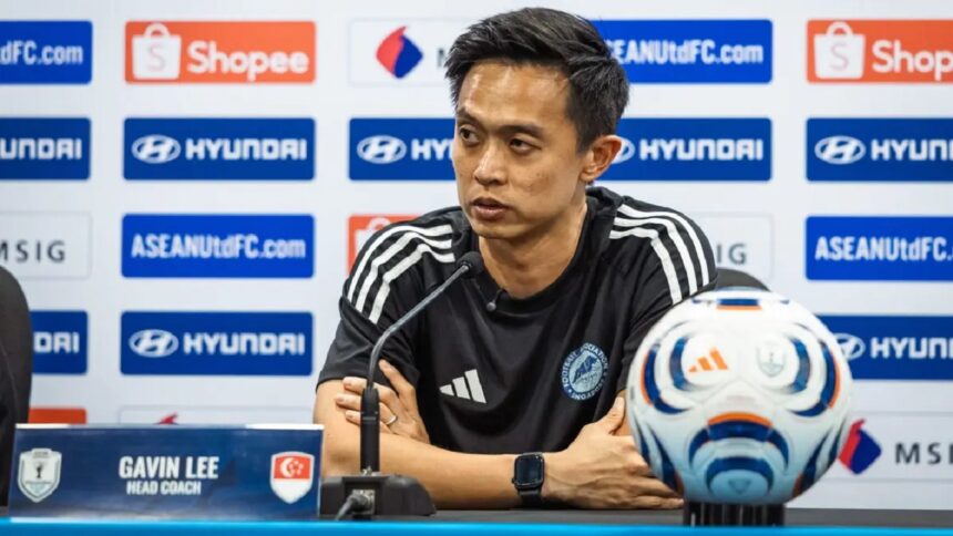 Press conference: man in black athletic shirt, nameplate reads Gavin Lee, head coach, at a football media briefing with logos in background