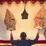 Performer on stage raises two ornate, kite-shaped puppets above their head in a traditional puppetry scene.