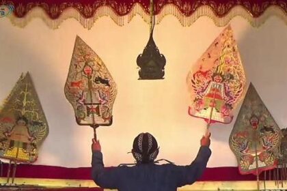 Performer on stage raises two ornate, kite-shaped puppets above their head in a traditional puppetry scene.