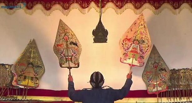Performer on stage raises two ornate, kite-shaped puppets above their head in a traditional puppetry scene.