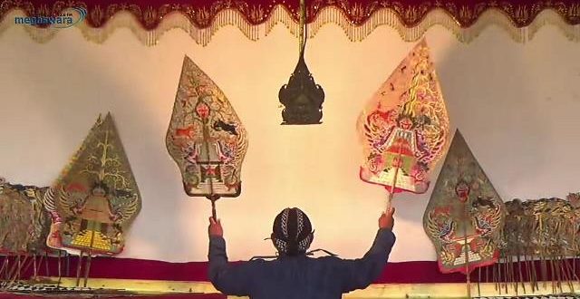 Performer on stage raises two ornate, kite-shaped puppets above their head in a traditional puppetry scene.