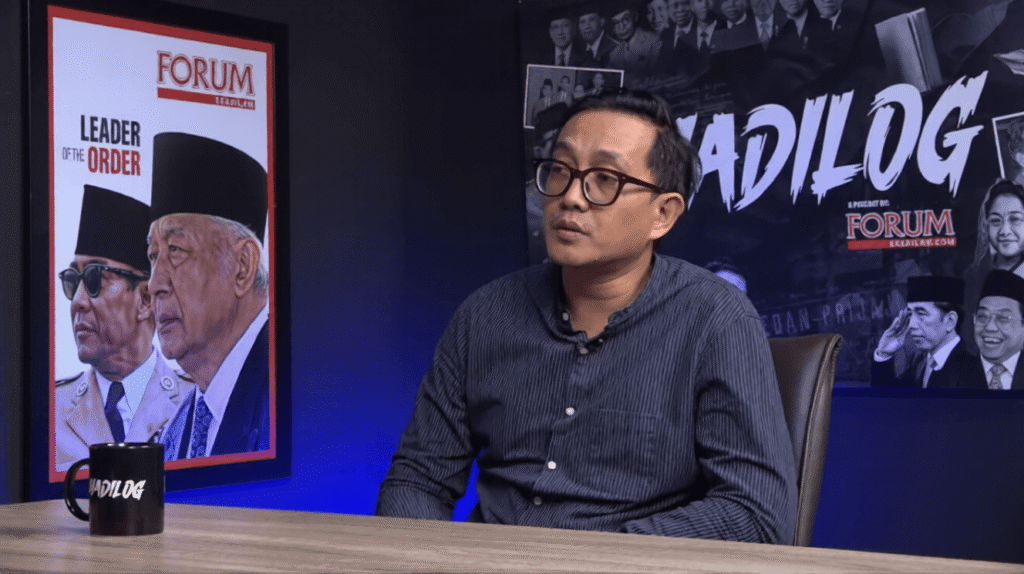 Man with glasses sits at a wooden desk in a studio interview set, blue lighting behind him, with posters that say FORUM and MADILOG on the wall and a MADILOG mug on the table.