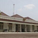 White colonial-style public building with a red-tiled roof, arched entrance, and a row of columns along a covered veranda; palm trees to the left.