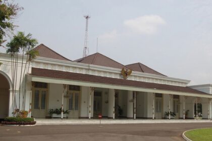 White colonial-style public building with a red-tiled roof, arched entrance, and a row of columns along a covered veranda; palm trees to the left.