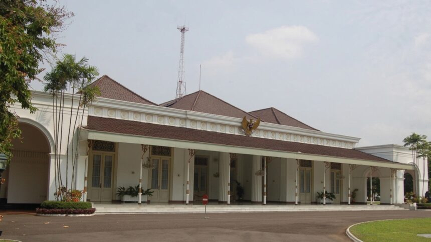 White colonial-style public building with a red-tiled roof, arched entrance, and a row of columns along a covered veranda; palm trees to the left.