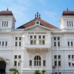 Front facade of Bank Indonesia building, a white neoclassical structure with red-tiled roof and decorative detailing under a blue sky