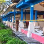 Officials in tan uniforms stand outside a blue-painted building under renovation, yellow caution tape blocking the entrance, debris nearby.