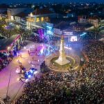 Large crowd gathers around a lit monument in a town square during an evening festival with a stage and colorful lights.