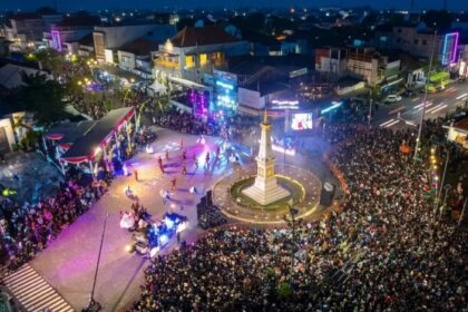 Large crowd gathers around a lit monument in a town square during an evening festival with a stage and colorful lights.