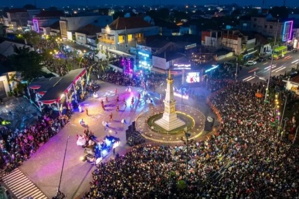 Large crowd gathers around a lit monument in a town square during an evening festival with a stage and colorful lights.