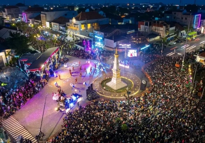 Large crowd gathers around a lit monument in a town square during an evening festival with a stage and colorful lights.