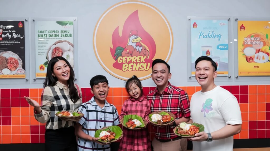 Five people smiling and posing with banana-leaf plates of food in a colorful restaurant, a large flame-logo with a chicken behind them.