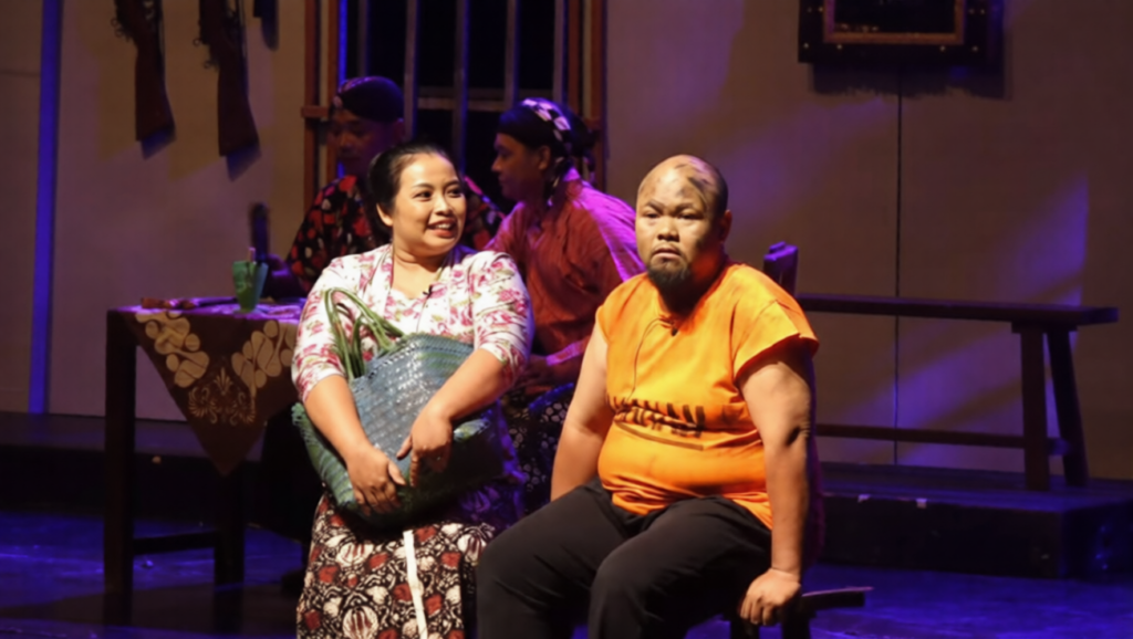 Ghost In Cell 2-ezremove - Mabur.co Two performers sit on a stage: a smiling woman with a woven bag, and a serious man in an orange shirt, during a theatre scene.