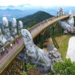 Pedestrian walkway held by giant stone hands, curving over a valley with travelers along a golden bridge.