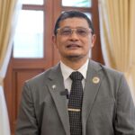 Man in a gray suit and tie standing indoors, wearing glasses and pins on lapel, in a formal room with beige curtains behind him.