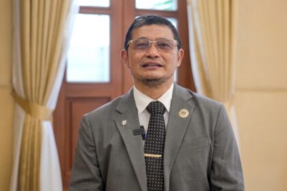 Man in a gray suit and tie standing indoors, wearing glasses and pins on lapel, in a formal room with beige curtains behind him.
