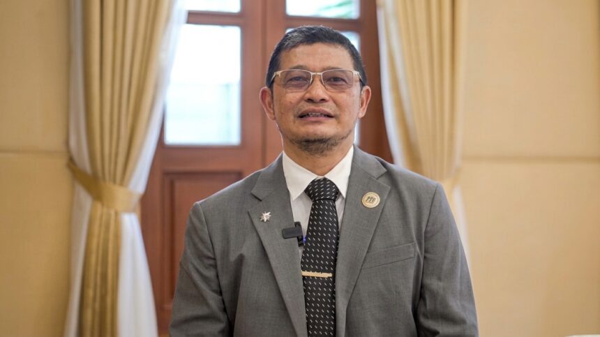 Man in a gray suit and tie standing indoors, wearing glasses and pins on lapel, in a formal room with beige curtains behind him.