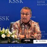 Middle-aged man in a batik-patterned shirt speaks at a panel, seated behind a table with a microphone, a flower arrangement to the left, and a blue backdrop reading 'KSSK'.
