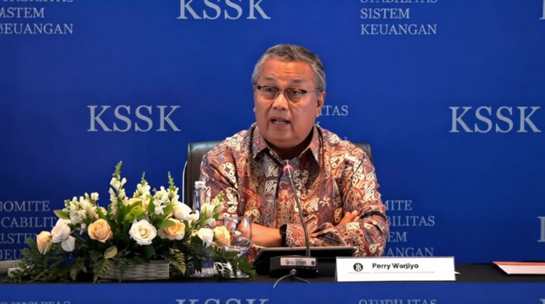 Middle-aged man in a batik-patterned shirt speaks at a panel, seated behind a table with a microphone, a flower arrangement to the left, and a blue backdrop reading 'KSSK'.