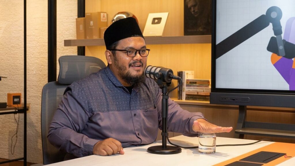 Guru Gembul by his own channel FIXX - Mabur.co Podcast host wearing a cap and glasses speaks into a microphone at a studio desk, with a glass of water nearby.