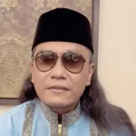 Man wearing a black songkok, sunglasses, and a light blue traditional outfit with patterned trim, seated indoors.