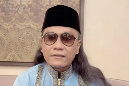 Man wearing a black songkok, sunglasses, and a light blue traditional outfit with patterned trim, seated indoors.