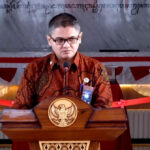 Man wearing batik shirt speaks at a wooden podium with a national emblem, flanked by flower arrangements at a formal event.